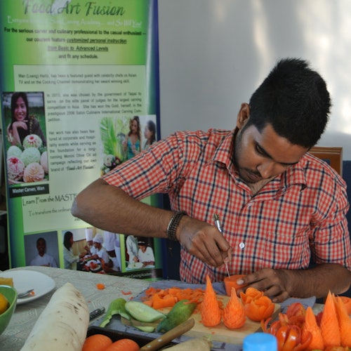 Un bărbat într-o cămașă cu carouri sculptează modele intricate în morcovi pe o masă încărcată cu diverse legume și fructe.