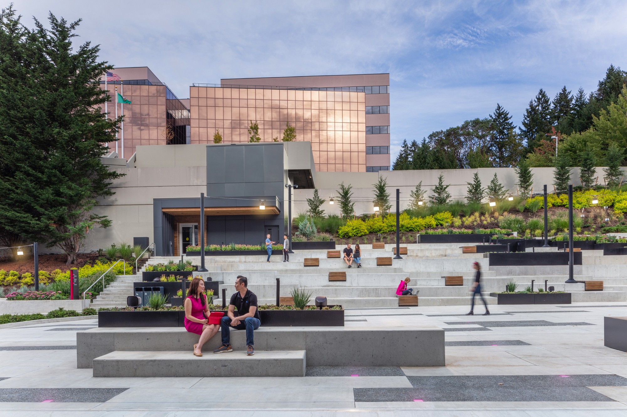 Two individuals seated on a concrete plinth, with the amphitheater adorned with wooden benches and planters visible beyond.