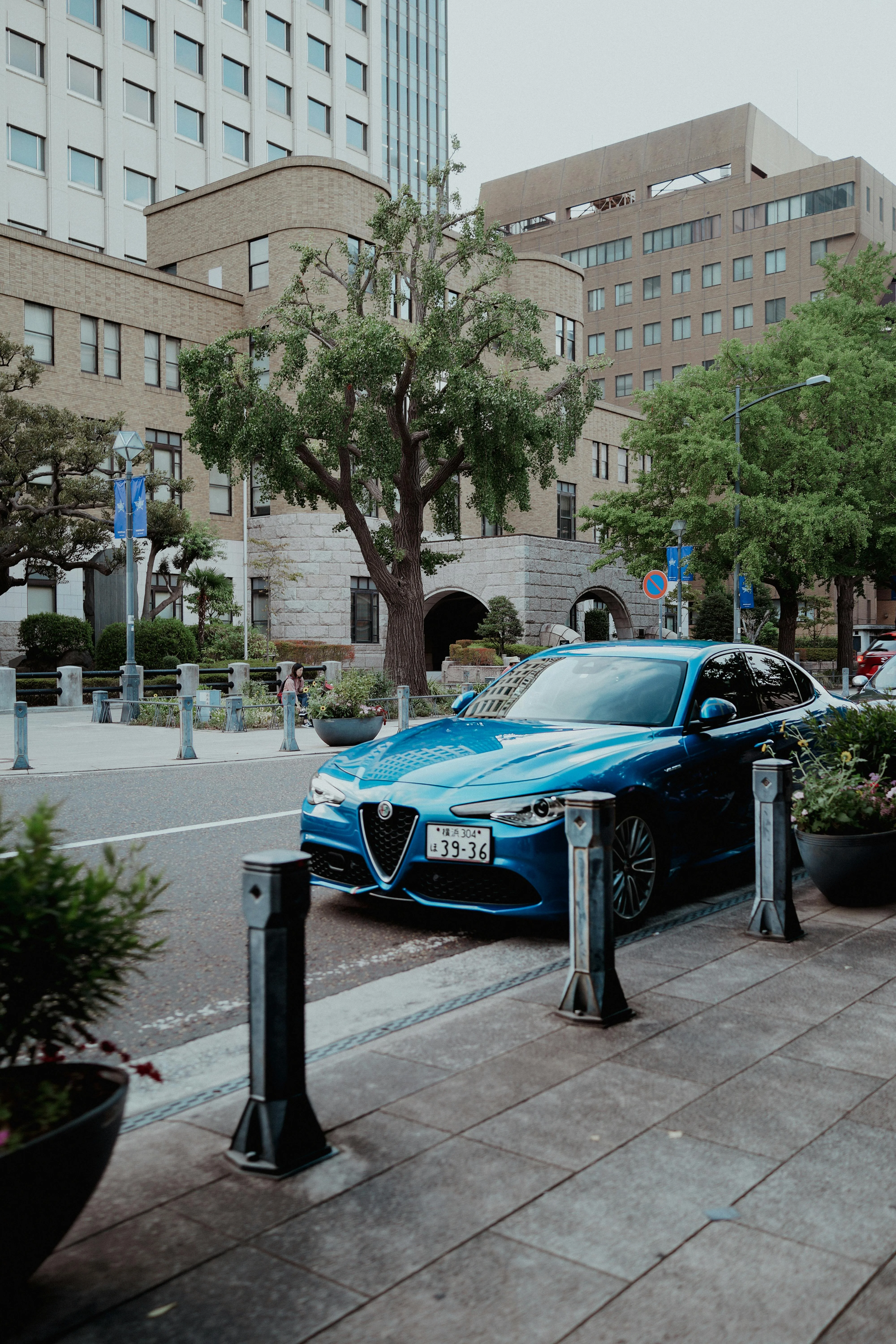 Une Alfa Romeo bleue stationnée dans une rue élégante, entourée de bâtiments modernes et d’arbres. La berline italienne se distingue par son design affûté, sa calandre triangulaire emblématique et son allure sportive, contrastant avec l’environnement urbain calme et structuré.