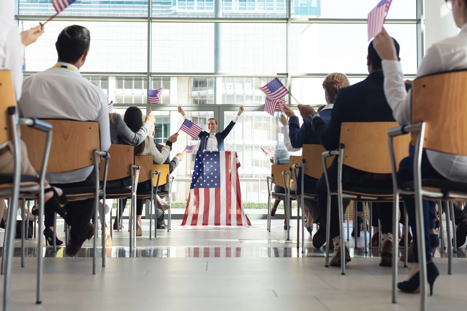 A woman in a dress stands before an audience, presenting with decor and flags in a bright indoor space.