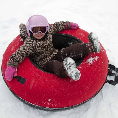 Child in a leopard print jacket, pink gloves, and a pink helmet sitting on a red snow tube in the snow, smiling.