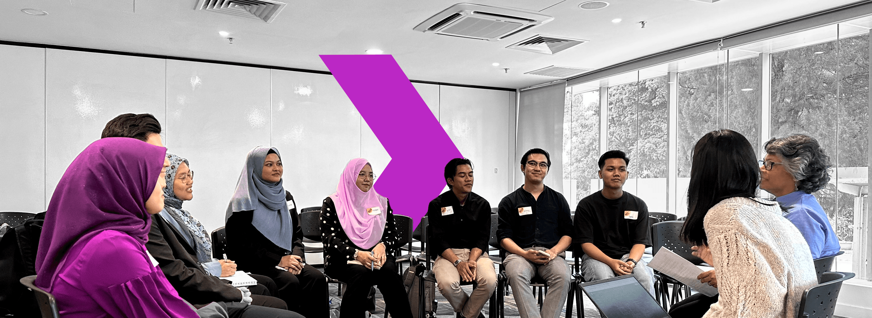A group of people in a workshop setting, seated in a circle, engaged in discussion with a large purple arrow design in the background.