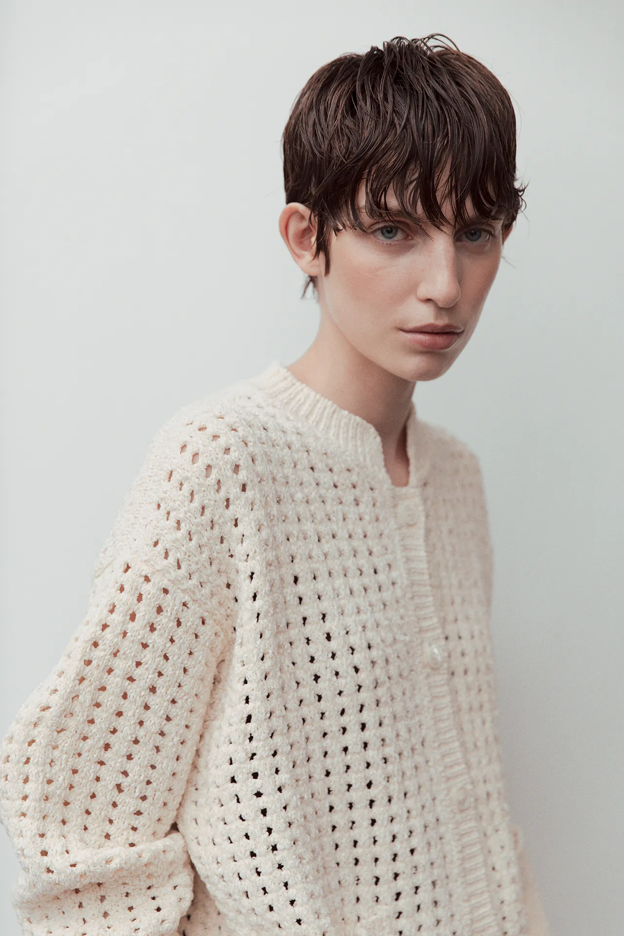 Rear view of a model from the Aida Barni SS26 collection wearing a cream chunky open-knit cardigan and brown wide-leg trousers standing near a white staircase.