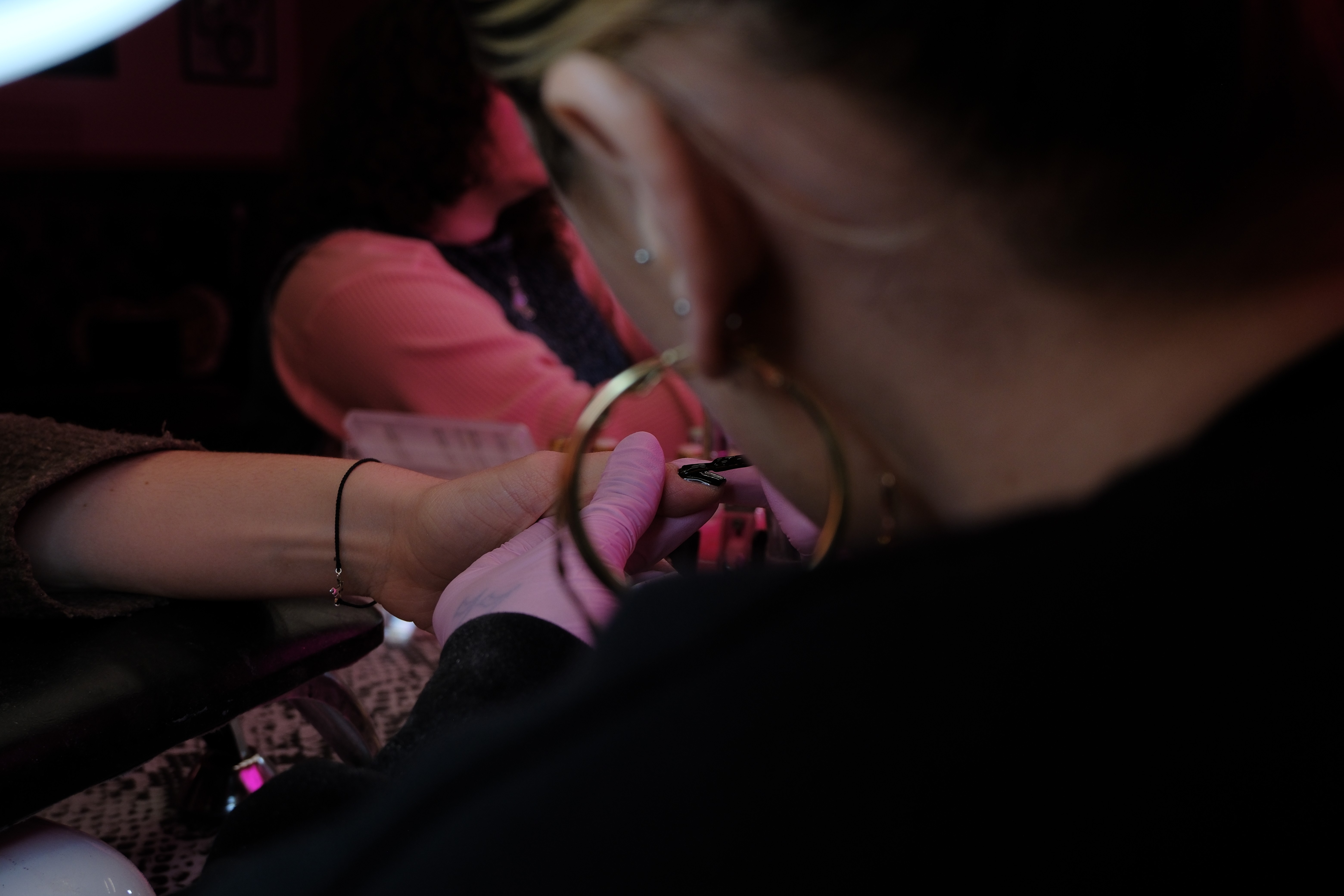 A focused shot over the shoulder of a technician (wearing large gold hoop earrings) as they work on a client’s nail under pink lighting.