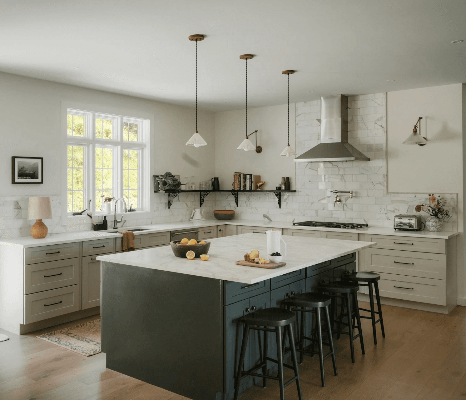 Modern white kitchen with gold hardware, marble island, wood stools, and windows overlooking a snowy forest.