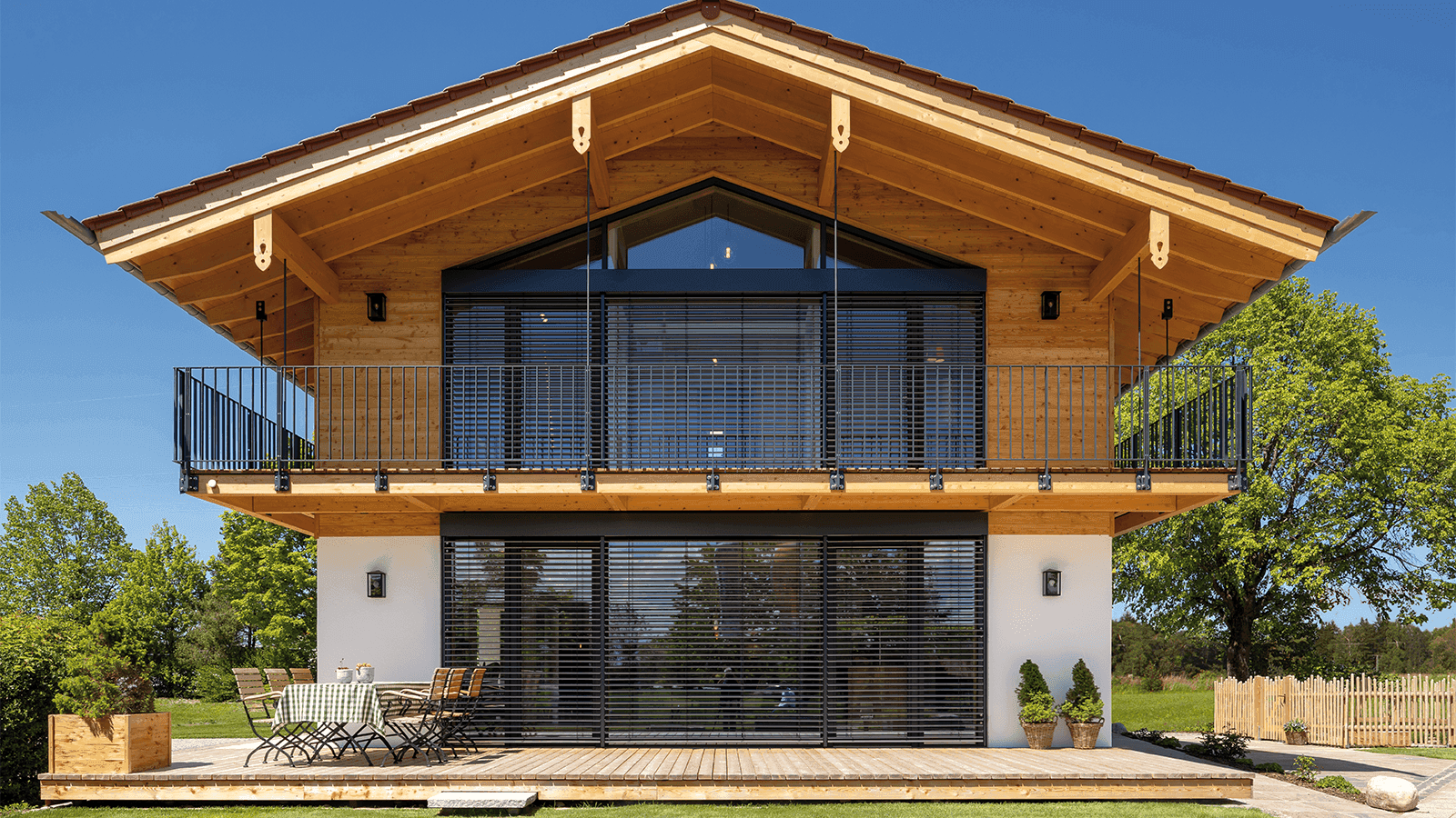 Modernes Holzhaus mit Balkon und Terrasse; Blick auf eine Gartensitzgruppe vor bodentiefen Fenstern.