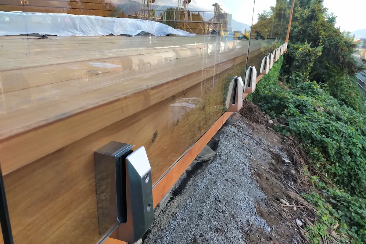 Close-up of frameless glass railing base clamps on wooden decking, showing precision hardware and installation detail by Tenmar.
