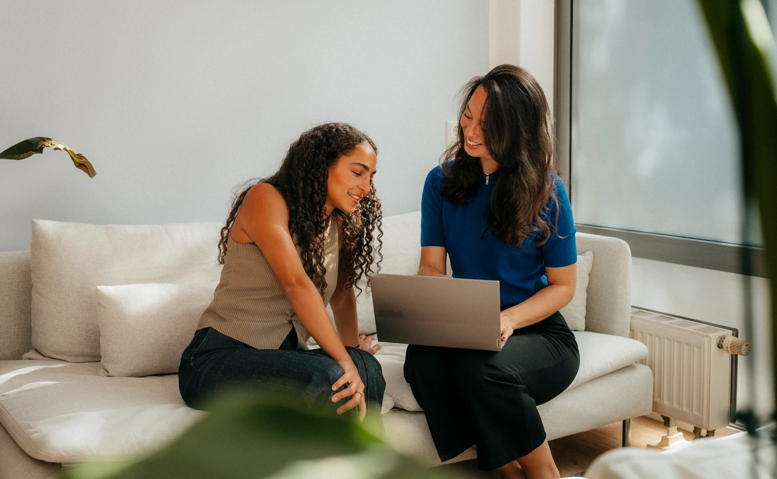 Zwei lächelnde Frauen sitzen gemeinsam auf einem Sofa und schauen auf einen Laptop