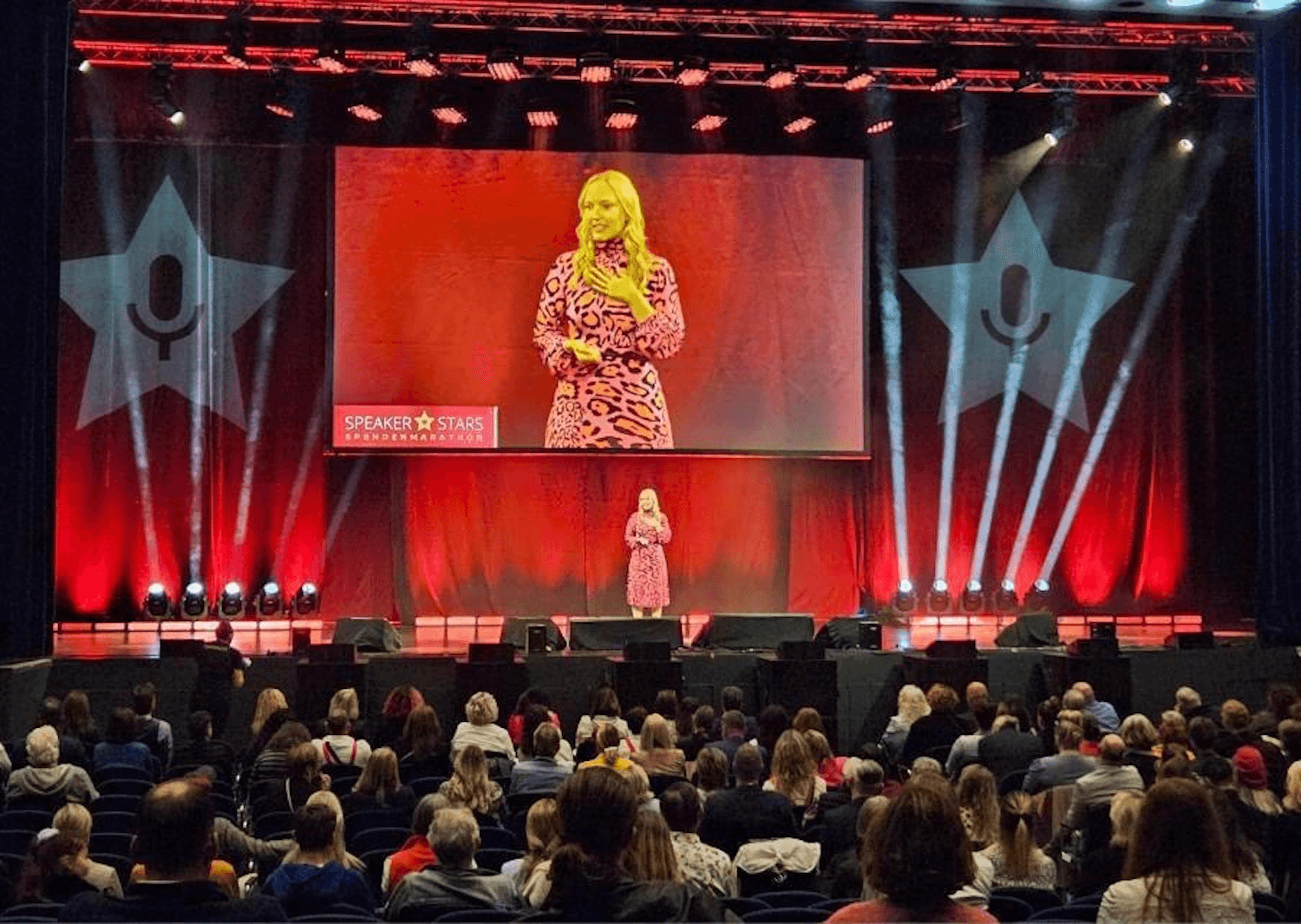 Simone Herzog speaking on a large illuminated stage before a seated audience.