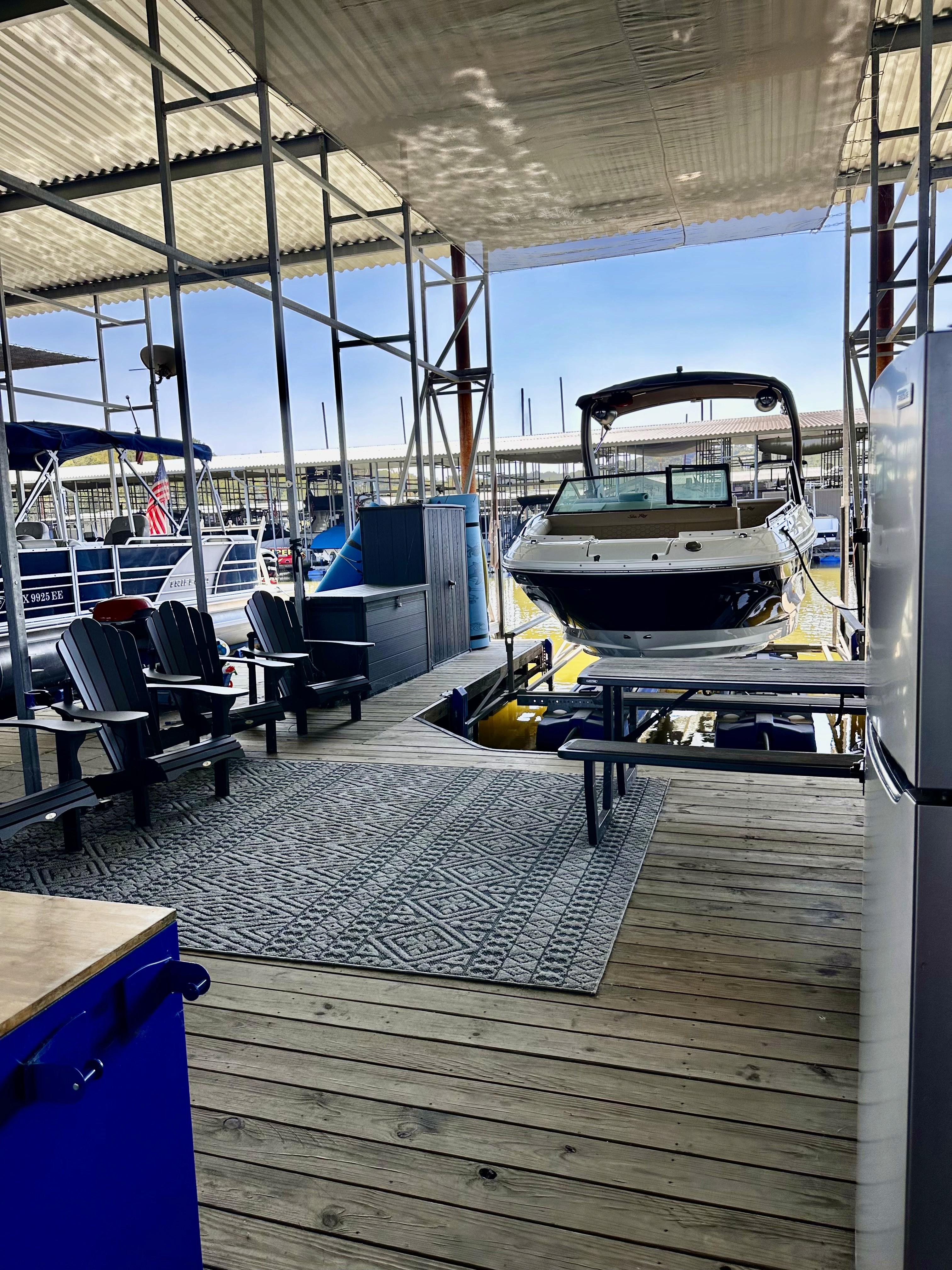 A boat is docked in a covered marina with wooden decking, surrounded by seating areas with Adirondack chairs and a patterned outdoor rug, set against a backdrop of other boats and blue skies.