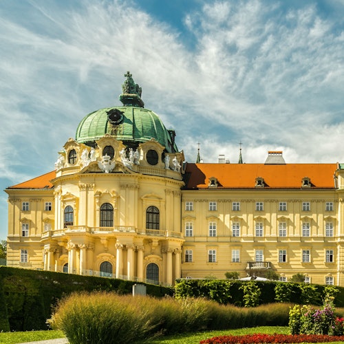 Ornate yellow and brown building with a green dome, classical architecture, and manicured gardens under a partly cloudy sky.