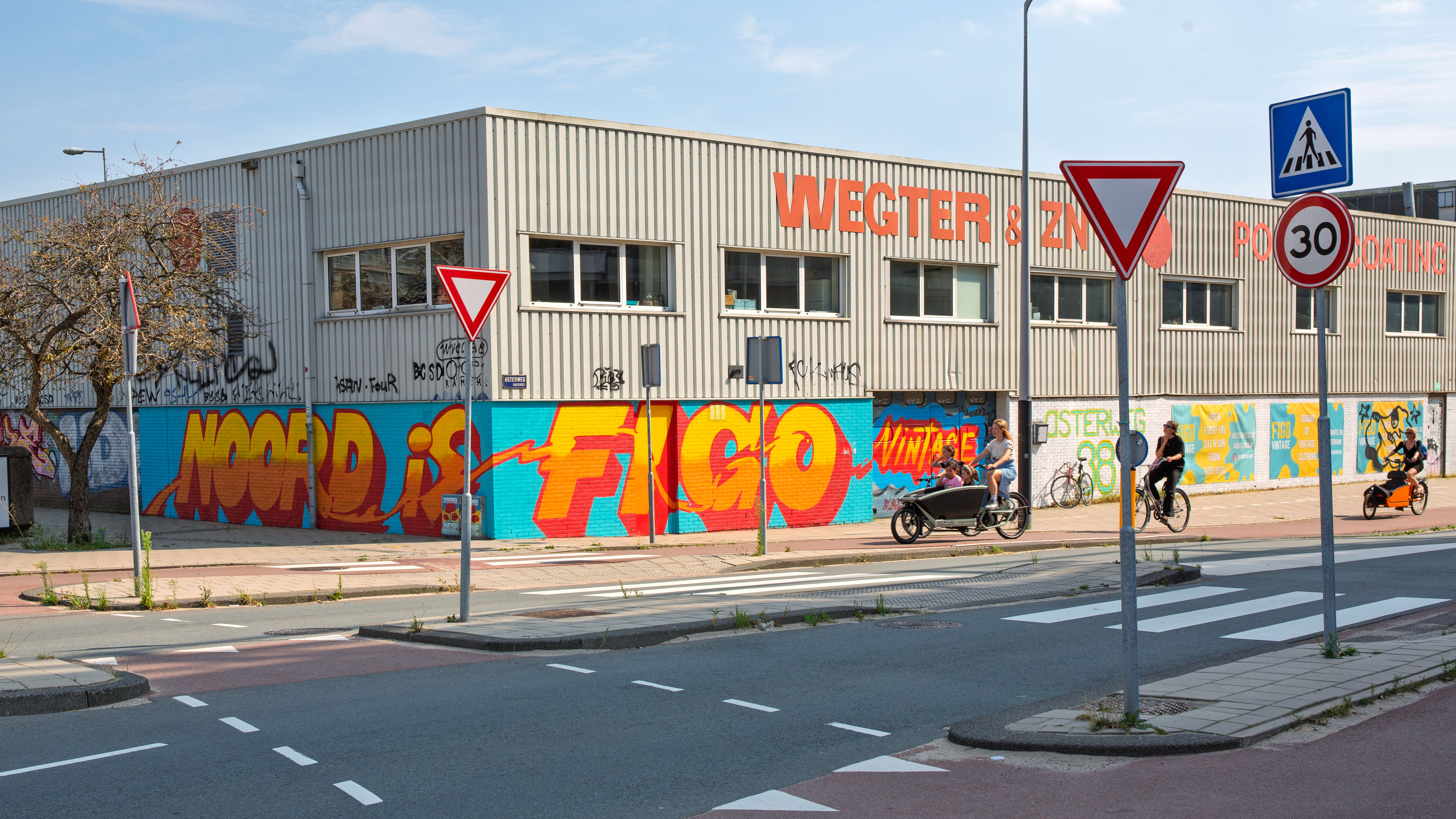 People cycling past a building with graffiti and street signs in the