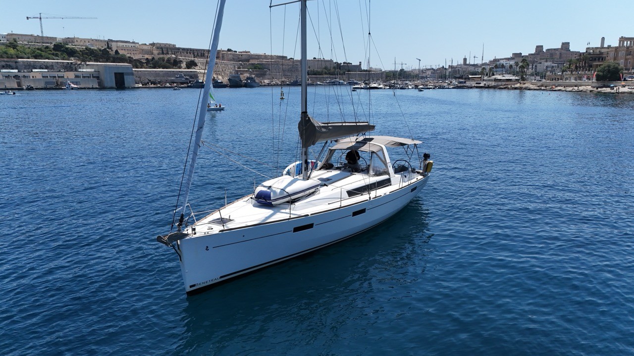 A Beneteau Oceanis 45 sailing yacht at anchor in a harbor, showing its long foredeck, furled headsail, and dinghy stowed on deck, with a fortified city skyline in the background.