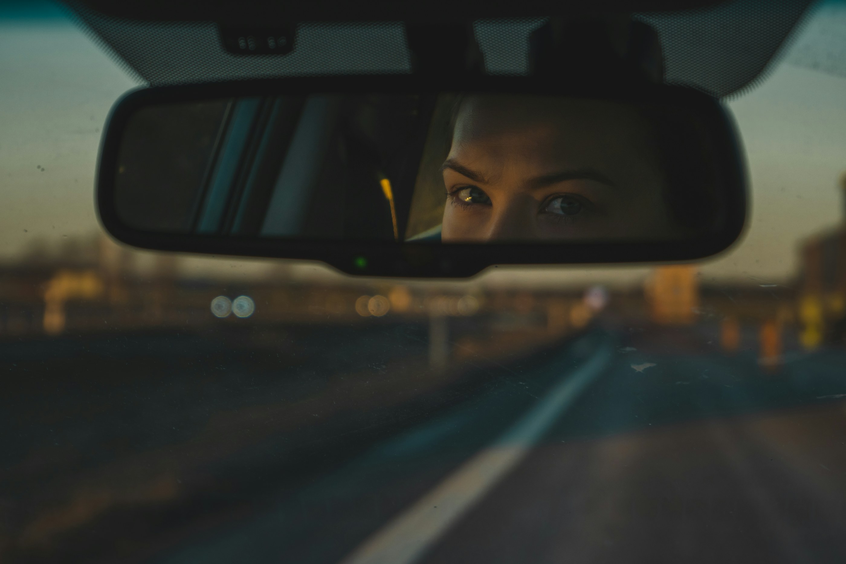 A driver's eyes reflected in the rear-view mirror on a highway at dusk