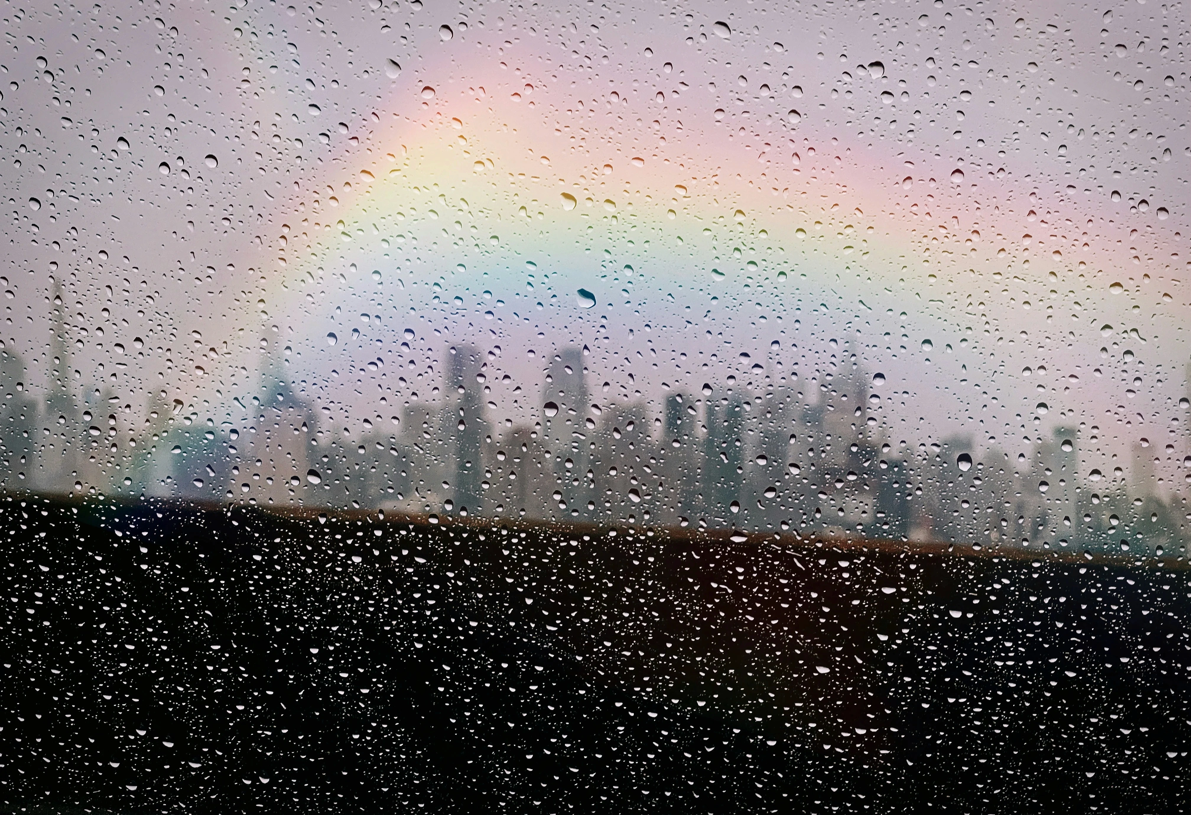Rainbow over the New York City skyline seen through a rain-covered window, symbolizing queer identity, hope, and belonging in an urban environment.