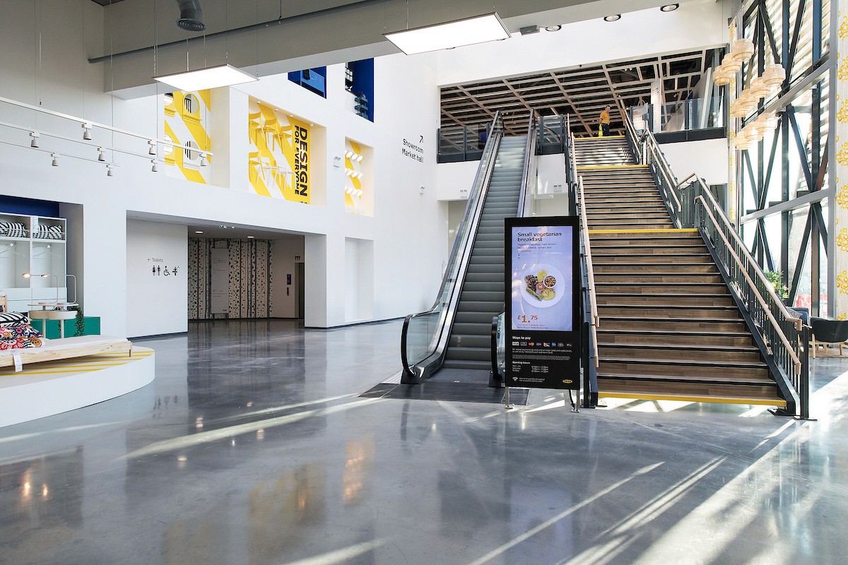 Interior of an IKEA store entrance featuring wooden stairs, an escalator, and digital signage. The minimalist architecture reflects the brand's 'Democratic Design' principles within a bright, high-ceiling commercial space.
