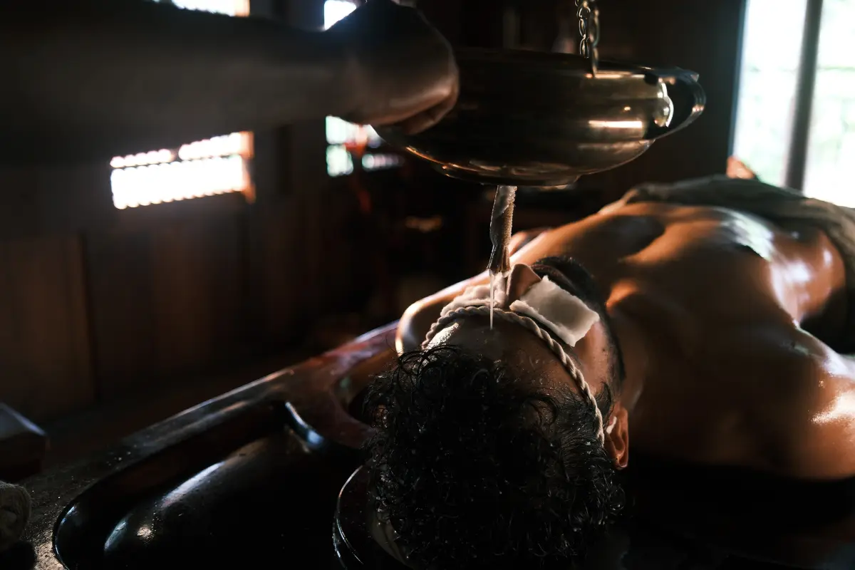 Traditional Shirodhara Ayurvedic oil treatment at Vedic Village Kodungallur, warm oil poured over the forehead in a wooden treatment room
