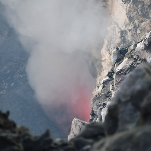 Uma paisagem rochosa e geotérmica com fumaça saindo de uma fissura, sugerindo atividade vulcânica. Um leve brilho vermelho é visível através da fumaça.