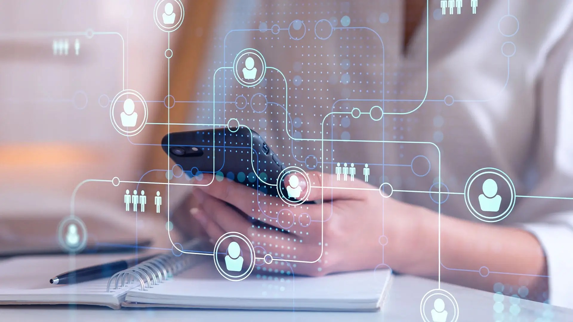 A person's hands holding a smartphone with a holographic display of social media and communication network icons, with a pen and notebook in the background.