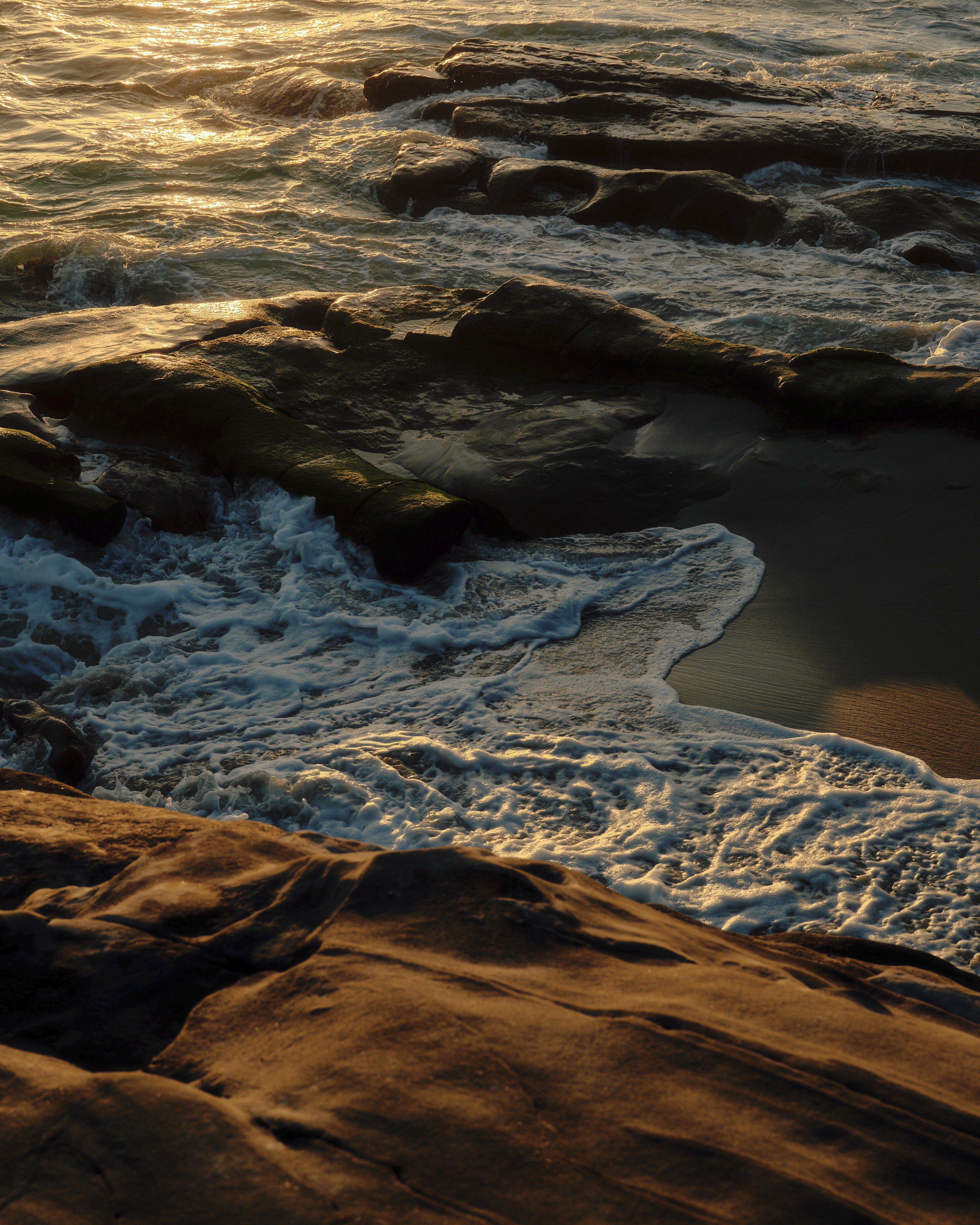 Ocean waves crashing against dark, moss-covered rocks on a beach at sunset, with golden light reflecting on the water