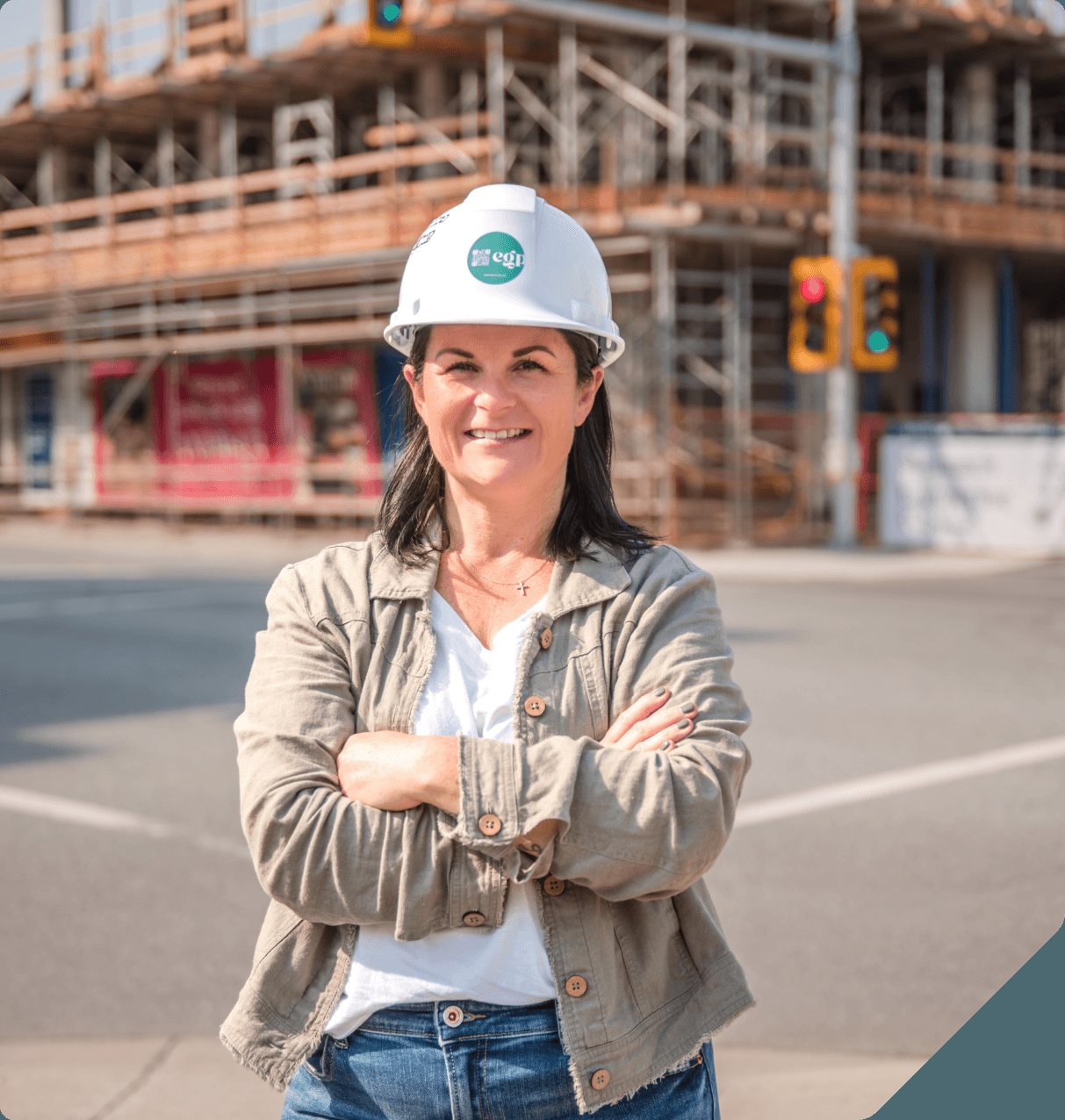 Carey Jones in a white hard hat and green jacket, arms crossed and smiling, in front of a building under construction