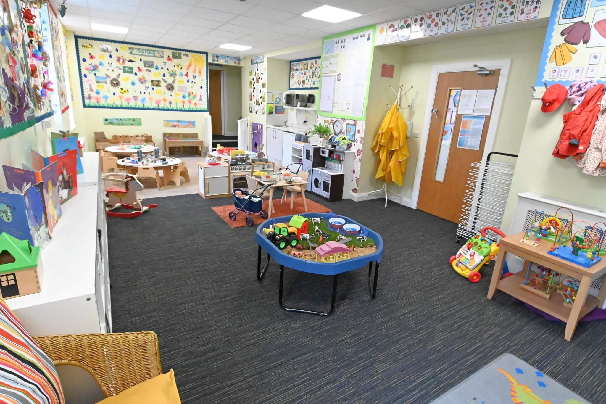 A colorful, well-equipped nursery classroom with a sand and water play table, kitchen play area, and child-sized furniture, ideal for early years education.