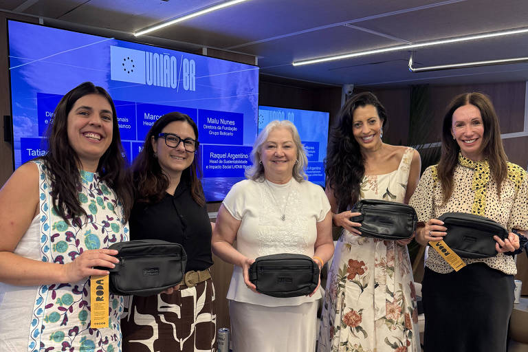 Cinco mulheres em pé em ambiente interno moderno seguram bolsas pretas. Elas vestem roupas claras e estampadas, com sorriso e postura alinhada. Ao fundo, tela azul com texto e iluminação de teto linear.
