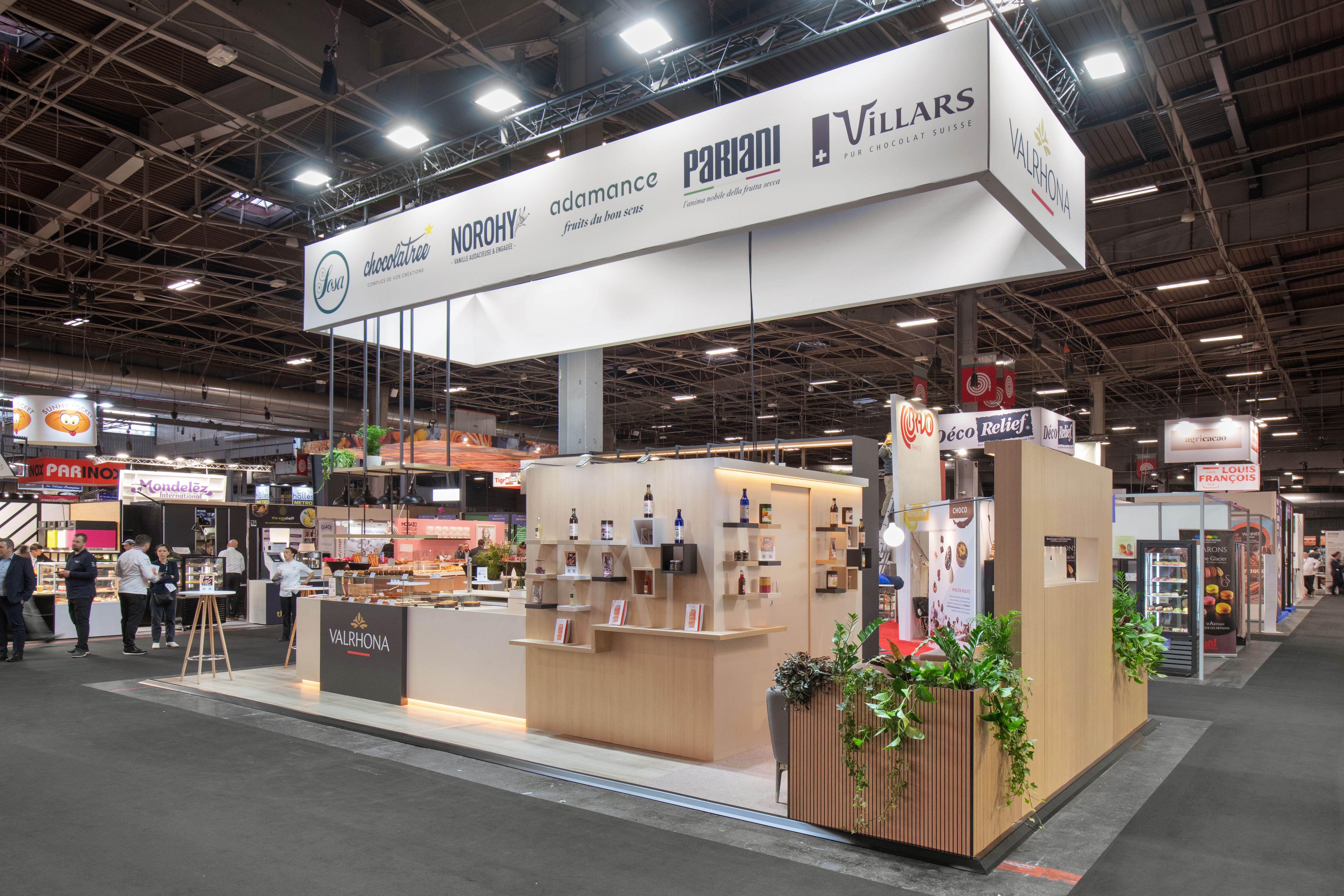 Stand Valrhona Bake and Snack 2026 conçu par Expace – vue trois quarts du stand avec aménagement sur-mesure bois clair, zone échange professionnels pâtissiers et chocolatiers et identité Valrhona