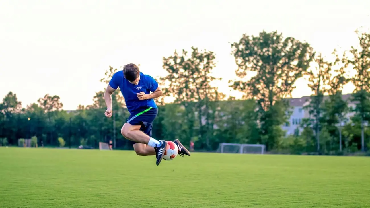 Das Bild zeigt einen Fußballspieler, der während eines Sprungs einen Trick ausführt, mit einem Ball in der Luft. Die Umgebung ist ein Fußballfeld im Freien, umgeben von Bäumen, mit einem sanften Abendlicht, das der Szene einen warmen Glanz verleiht. Der Athlet, der in einem blauen Trainingsshirt und Shorts gekleidet ist, führt die Bewegung mit Präzision aus, was dynamische Bewegung und Technikfokus widerspiegelt. Im Hintergrund sind ein leicht verschwommenes Feld und Torpfosten zu sehen, die die Aktion des Spielers als zentralen Fokus des Bildes betonen.