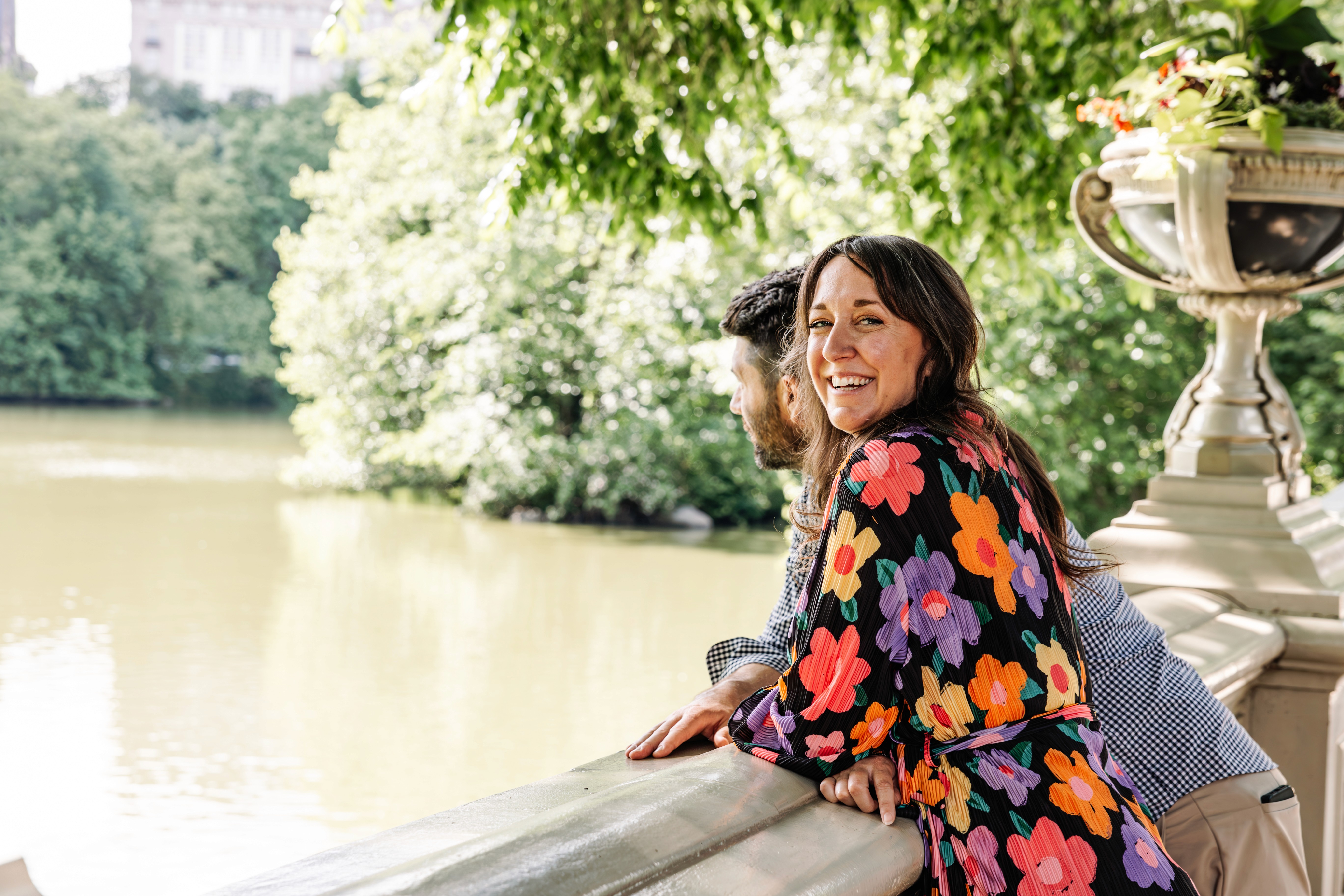 Woman in a bold floral dress laughing and leaning on the stone railing at Bow Bridge in Central Park, her partner close behind — Lizz Spano Photography, New York City couples photographer