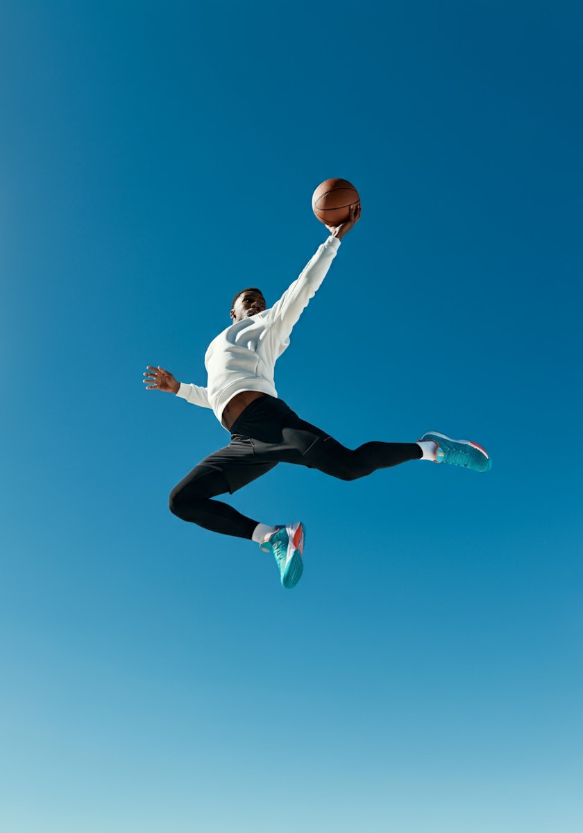 Athlete in mid-air leap with basketball, wearing white hoodie and turquoise sneakers against a clear blue sky.