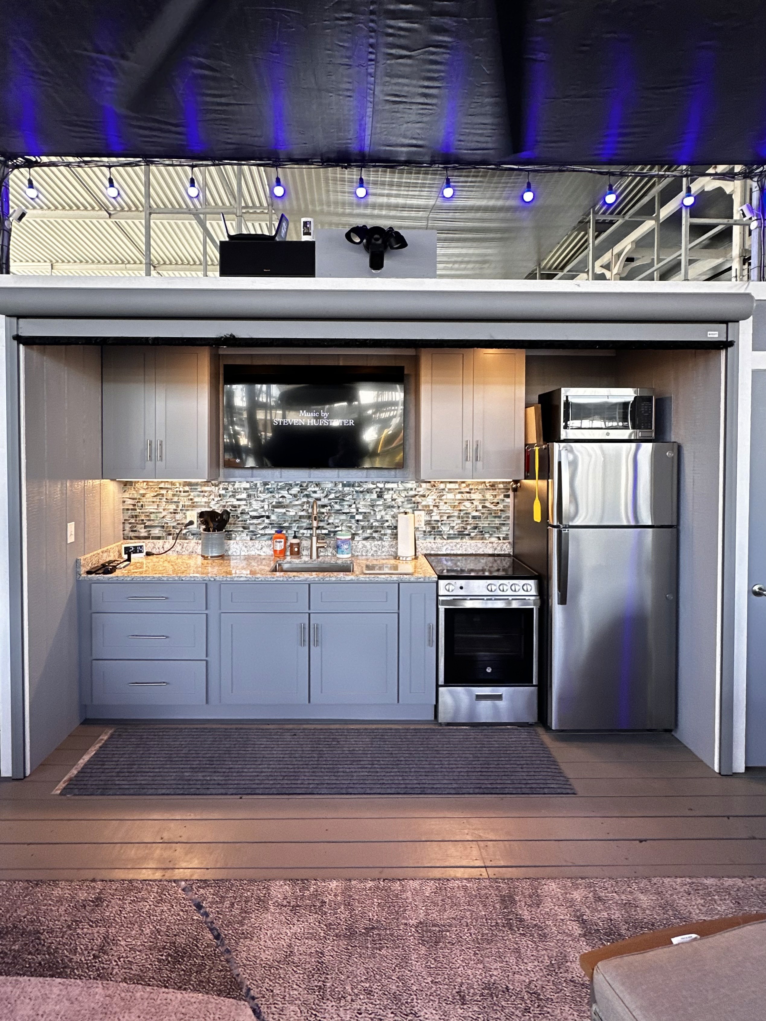 A modern, compact kitchen features sleek gray cabinetry, a stainless steel refrigerator and oven, and a backsplash with a mosaic tile design, illuminated by blue accent lighting.