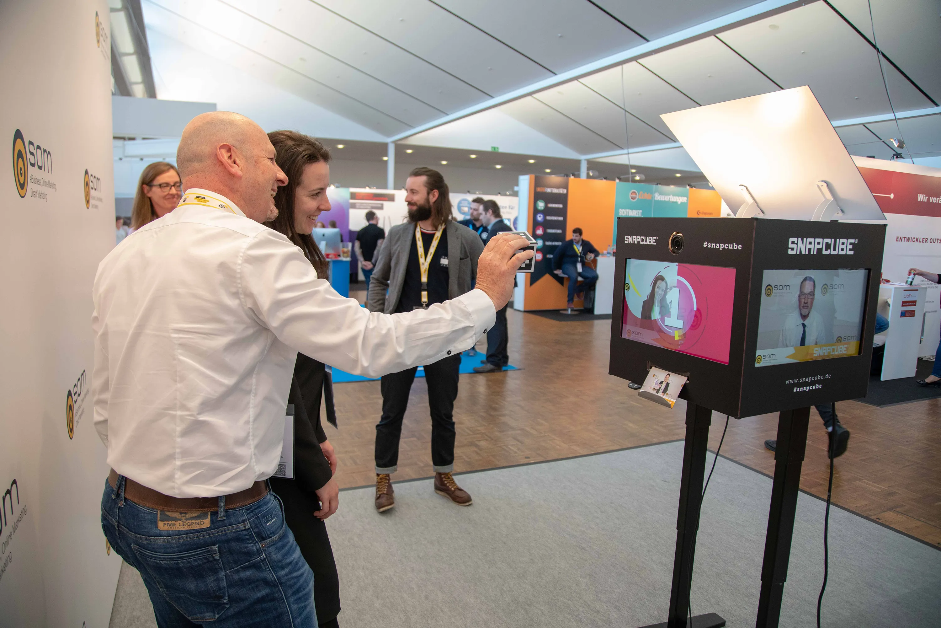 SnapCube Fotobox im Einsatz auf einer Messe: Zwei Besucher fotografieren sich DSGVO konform am CUBE mit vier Bildschirmen