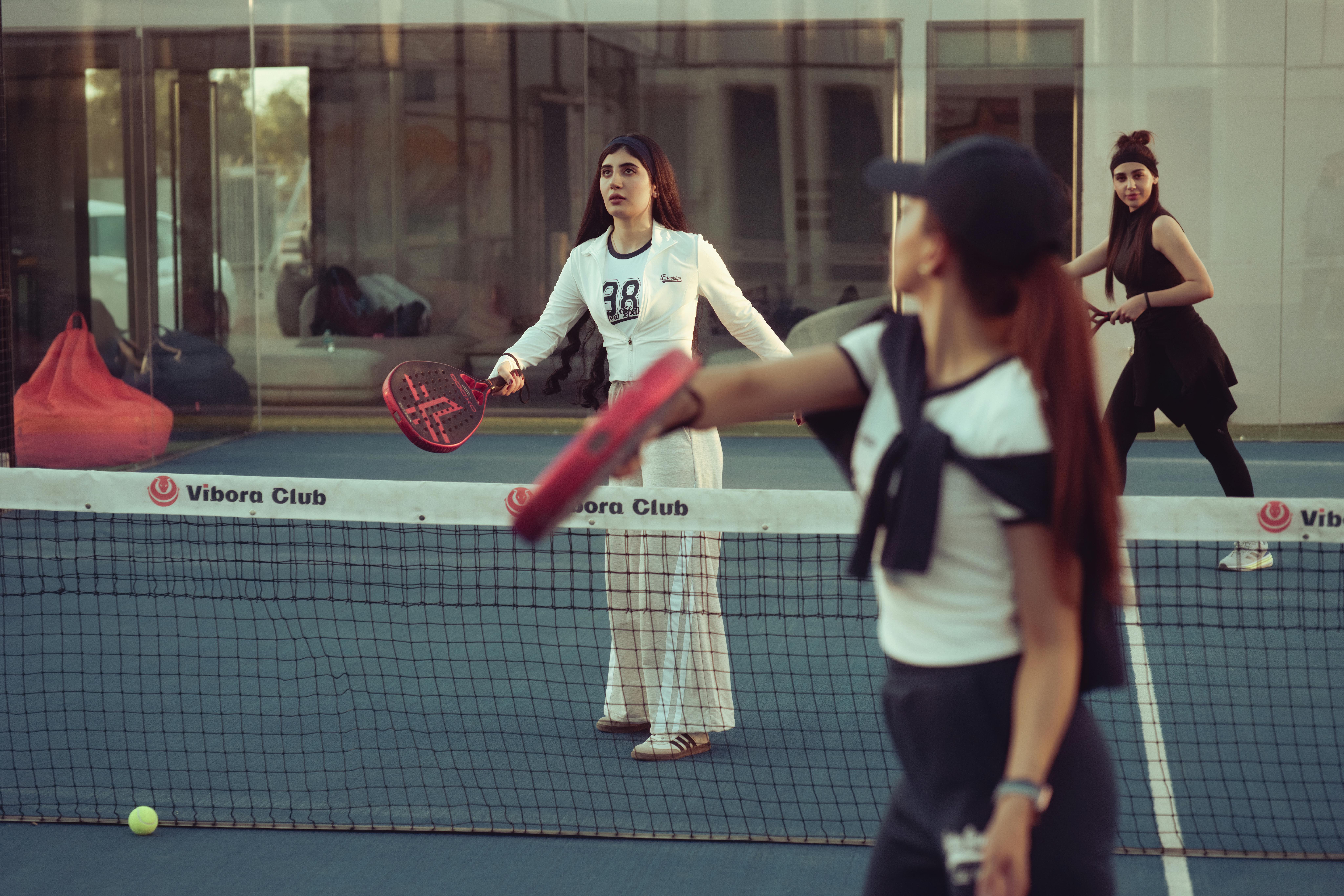 Women engage in a friendly paddle tennis match on an indoor court, with one serving the ball while another prepares to return, highlighting the energy and focus of the sport.