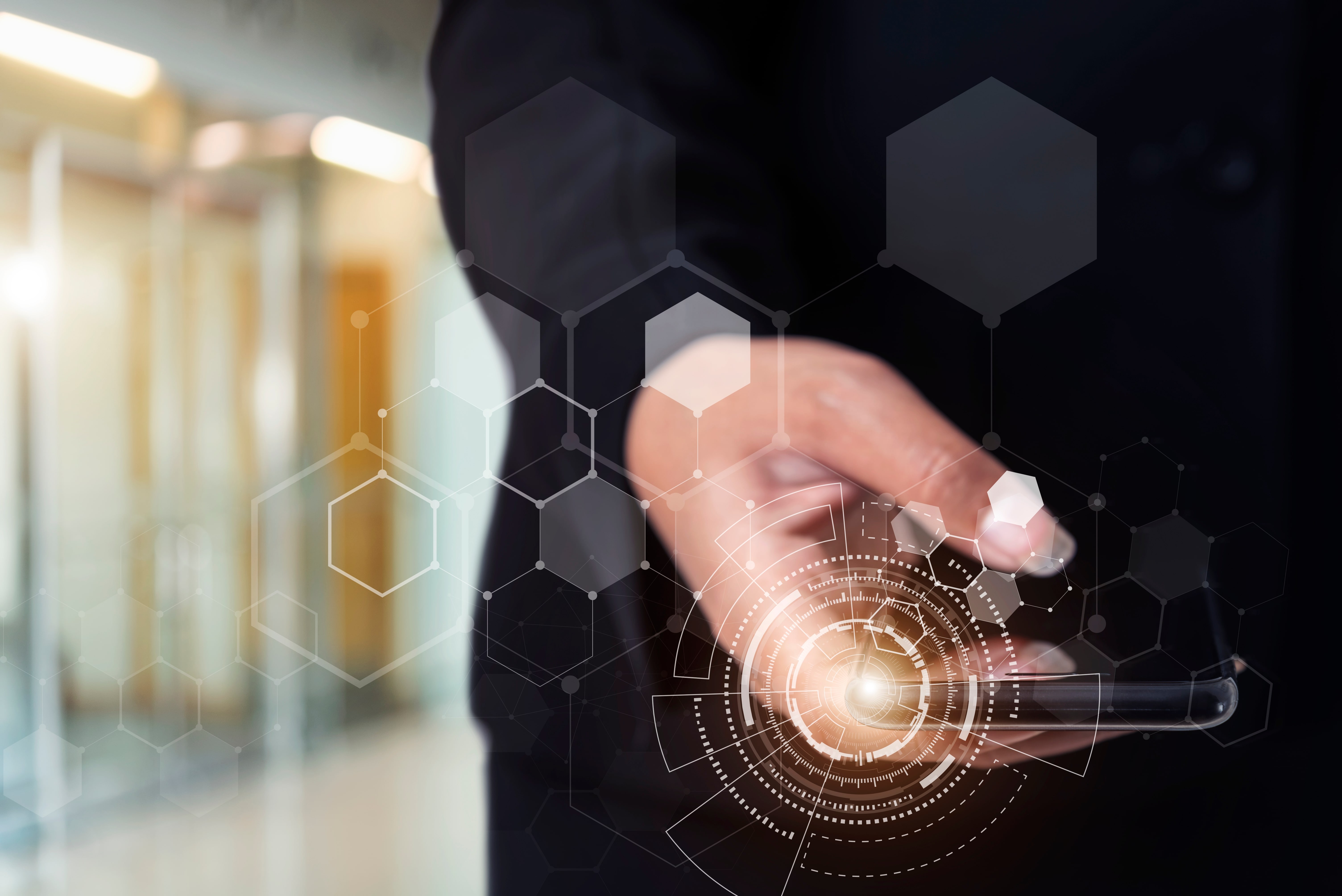 A conceptual image of a business professional in a suit holding a smartphone, with a glowing, futuristic digital interface and a hexagonal network graphic projected over their hand. This represents innovation, business technology, big data, cybersecurity, and digital transformation in a modern office environment