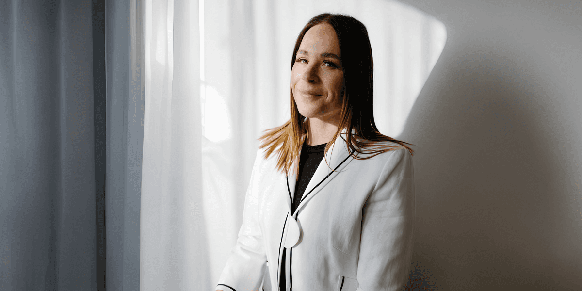 Professional woman in a white blazer standing by a sunlit window, smiling confidently in soft natural light.