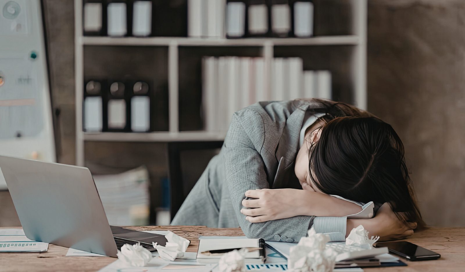 A tired small business owner resting her head on her desk beside a laptop and crumpled paper, frustrated from trying to build her own website.