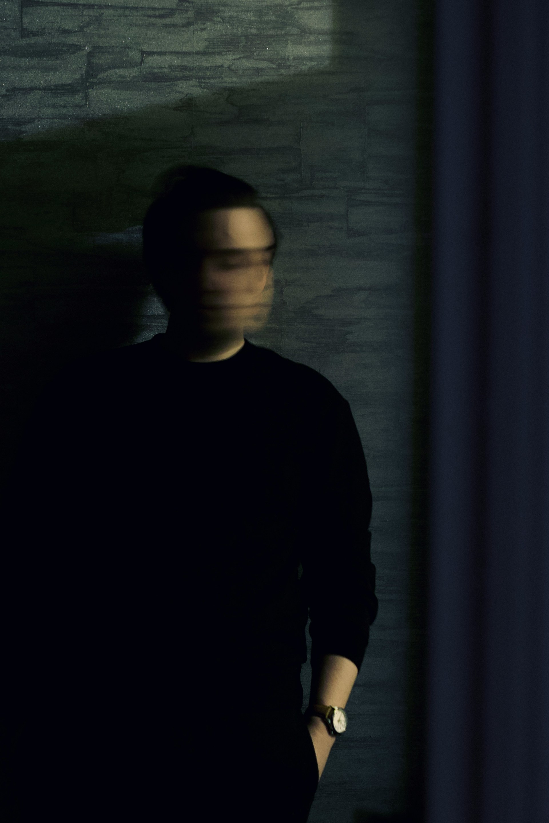 A blurred figure in dark clothing stands against a textured gray wall with one hand in their pocket, creating a mysterious and shadowy atmosphere.