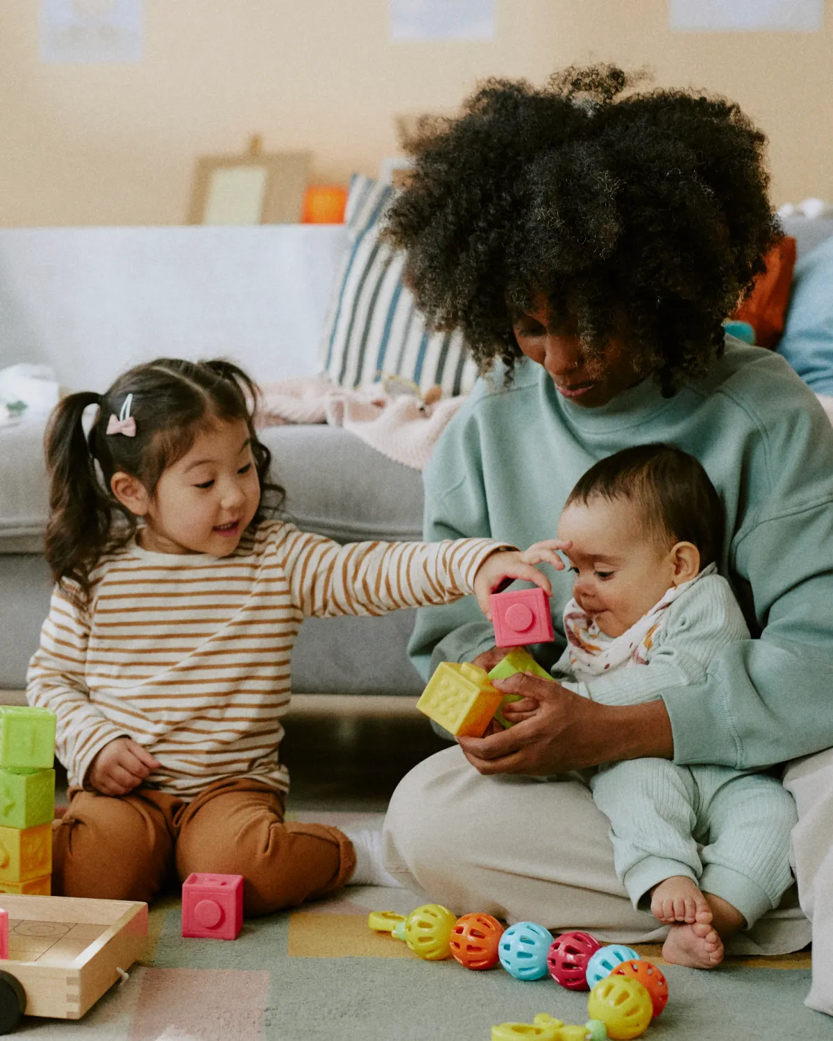 A caregiver sitting on the floor playing with a toddler and a baby using colorful building blocks