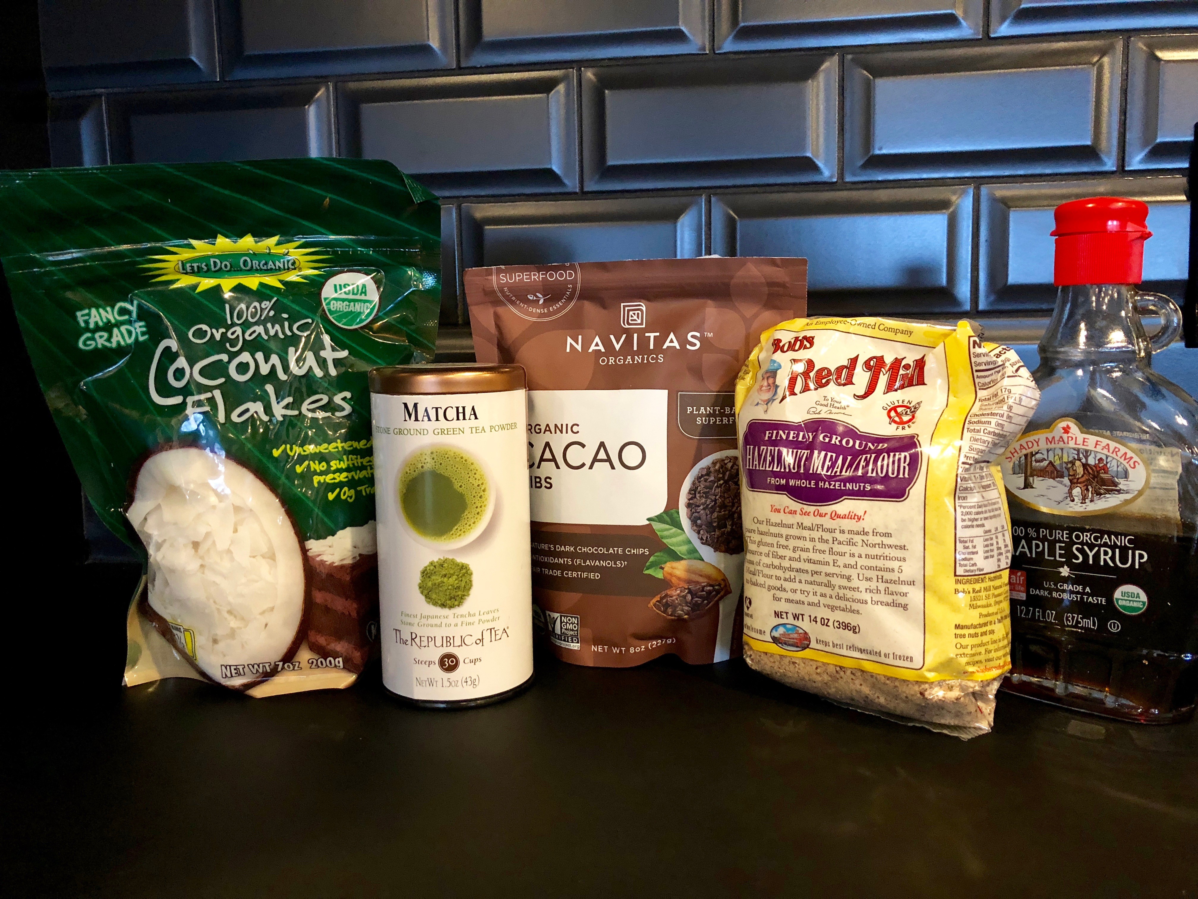 Organic food products displayed on a kitchen counter, including coconut flakes, matcha tea, cacao nibs, a grain mix, and a bottle of maple syrup, set against a textured black backsplash.