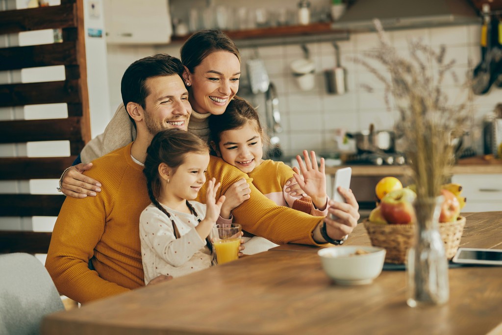 The image shows a happy family of four—two parents and two young daughters—taking a selfie together at their kitchen table. They are all smiling and waving at the camera, capturing a joyful moment. The kitchen setting is cozy and inviting, with a bowl of fruit and a glass of orange juice on the table, enhancing the warm, family-friendly atmosphere.