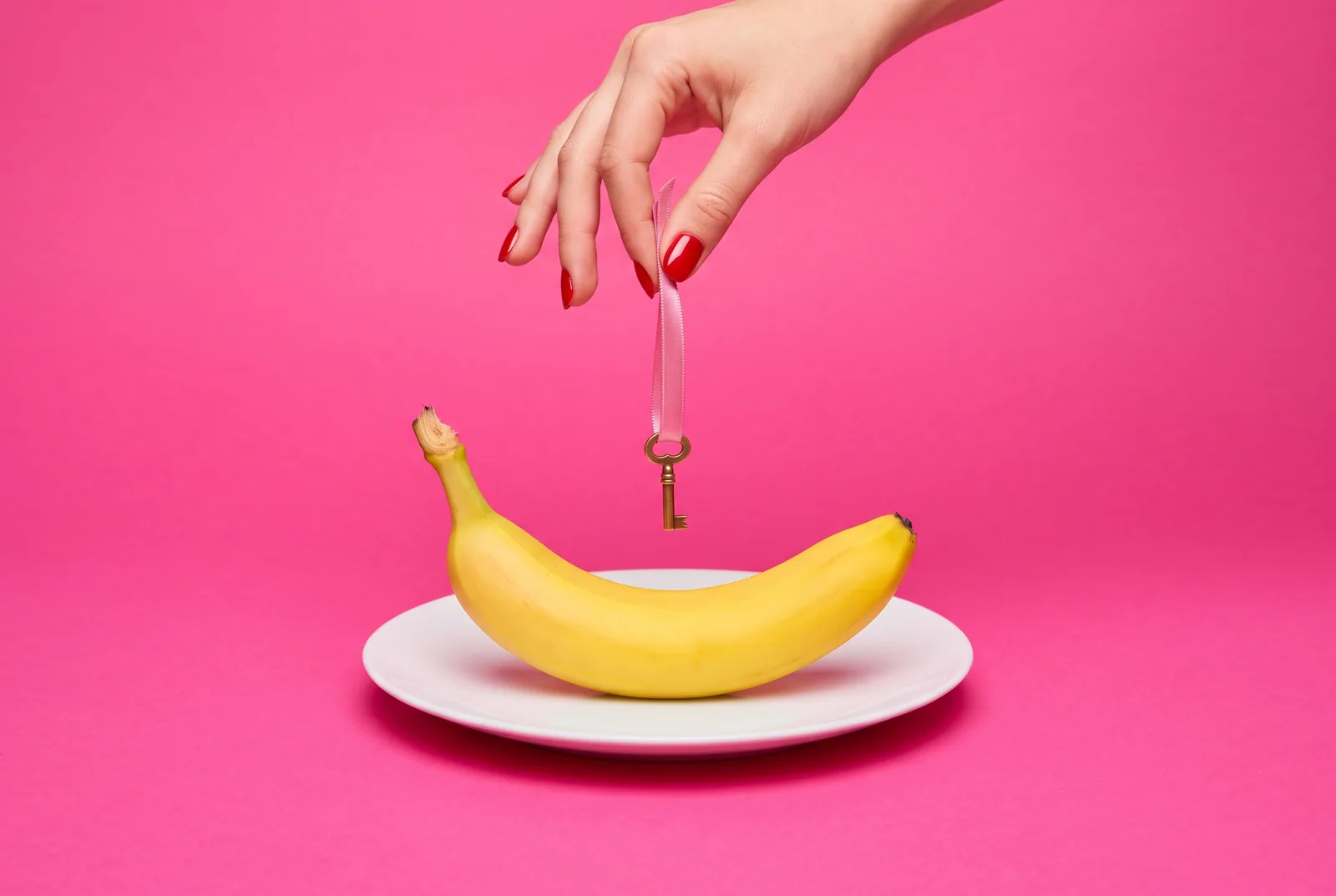 A hand with red nails holding a tiny brass key on a ribbon above a banana, a trusted person holding the Mac Screen Time passcode