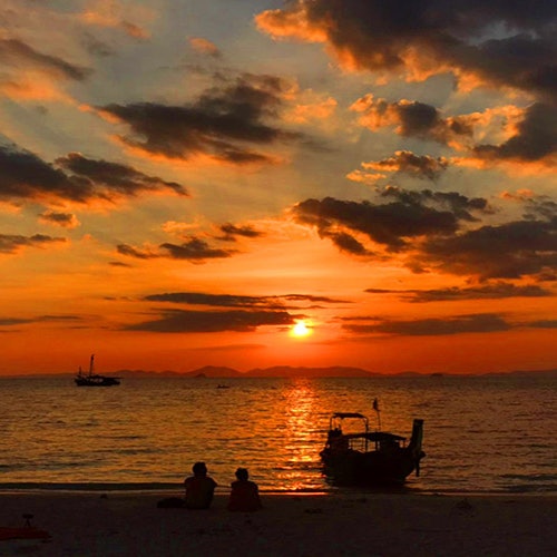 Two people sit on the beach, watching a sunset over the ocean with boats silhouetted against a colorful sky.