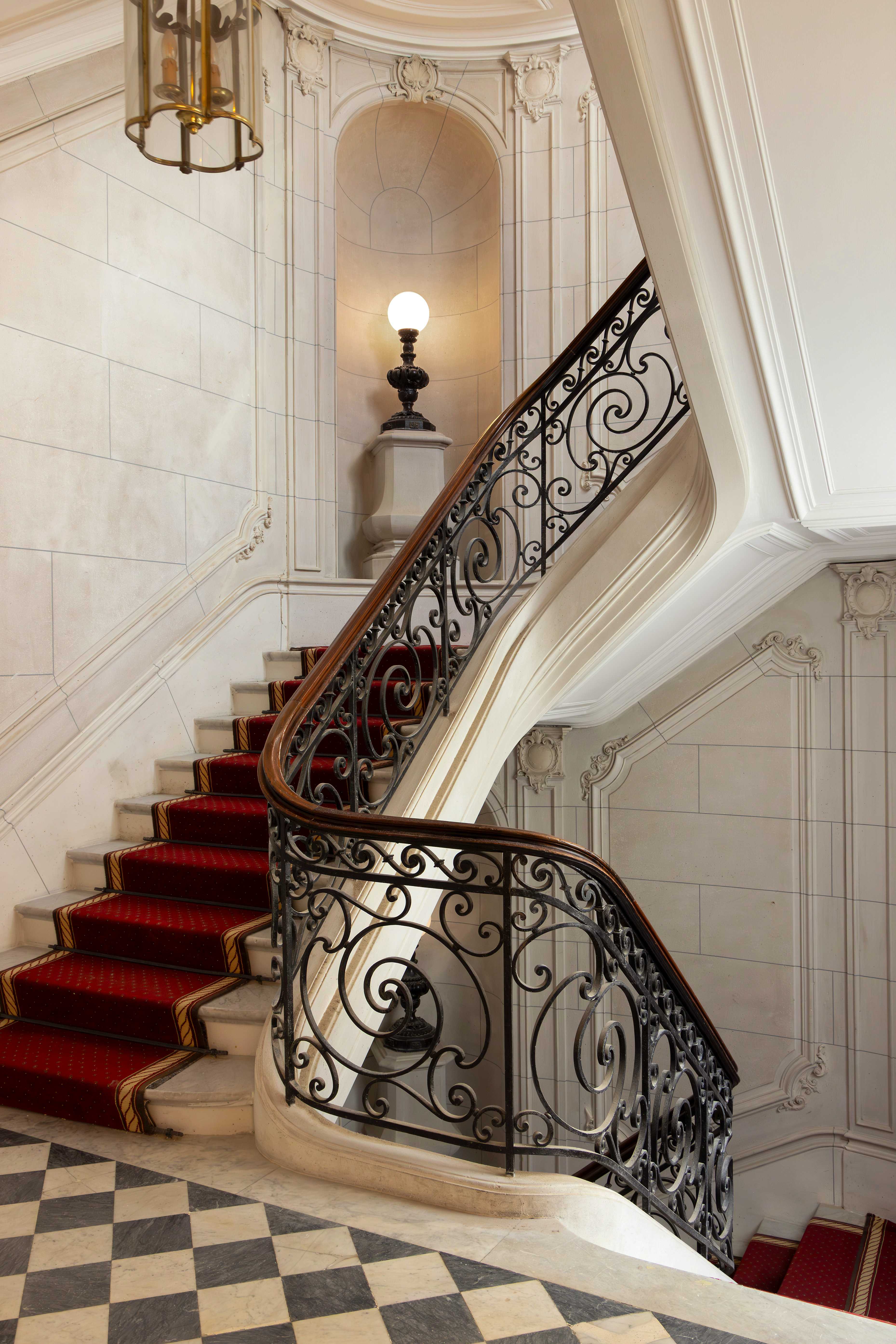 Escalier monumental en pierre avec rampe en fer forge ornee, tapis rouge, lanterne et niche avec lampe a globe