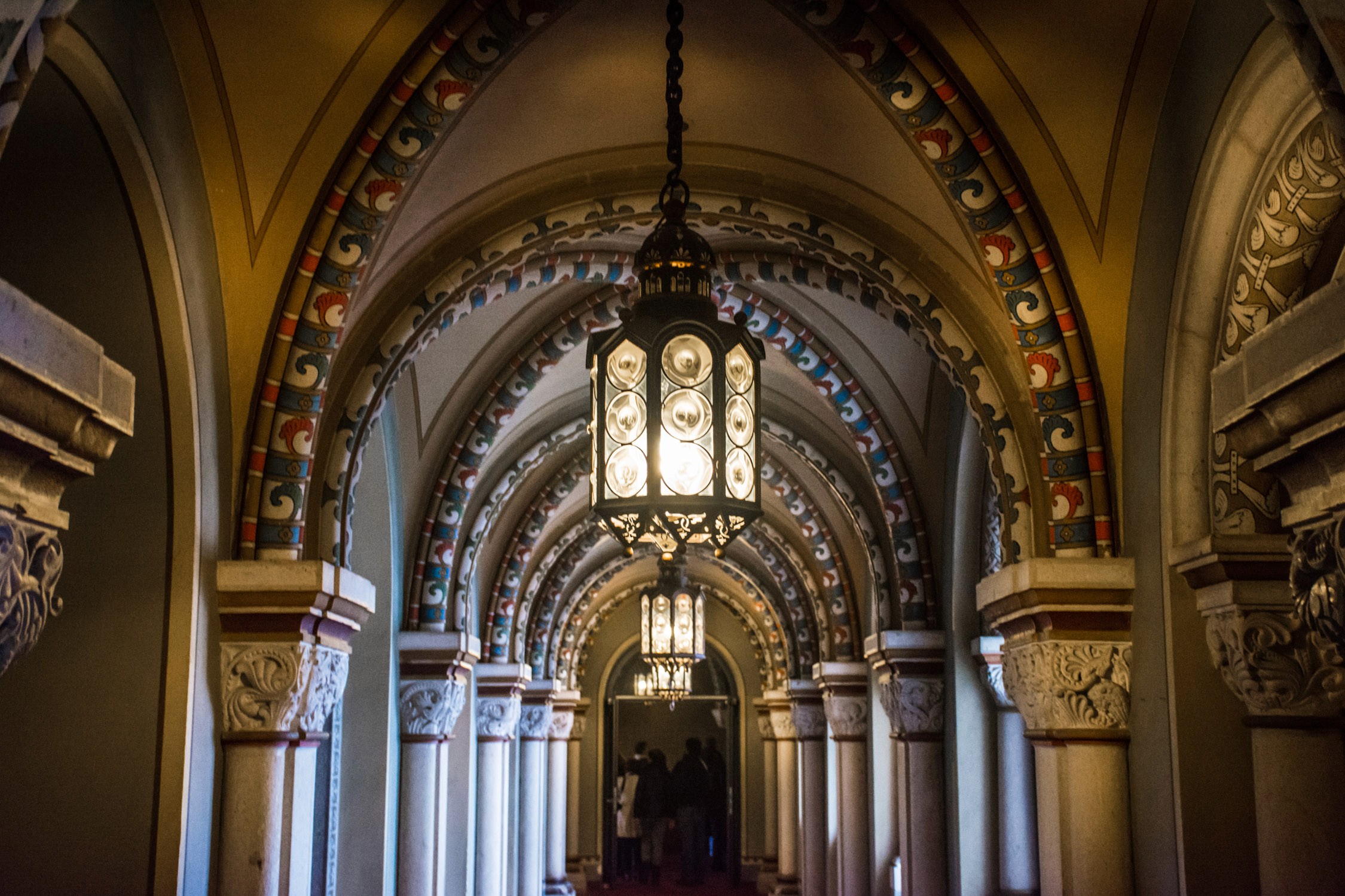 A long, arched stone corridor with ornate carvings and a large hanging lantern illuminates a distant altar in a church.