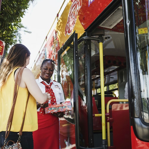 A woman in a yellow top boards a double-decker bus, greeted by a smiling conductor in a red and white uniform.