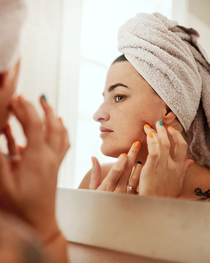 A woman with a towel on her head applying skincare in front of a bathroom mirror