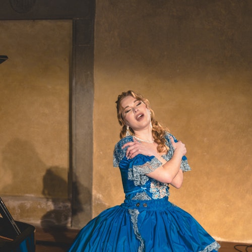 一位身穿蓝色复古风连衣裙的女子在昏暗的房间里摆出富有戏剧感的姿势，旁边可见一架钢琴的一部分。