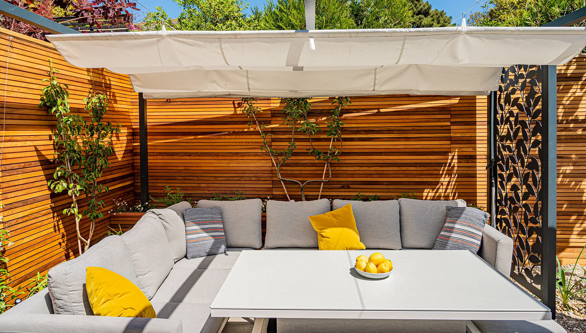 A cozy outdoor seating area with a gray couch, yellow cushions, and a white table under a shaded pergola.