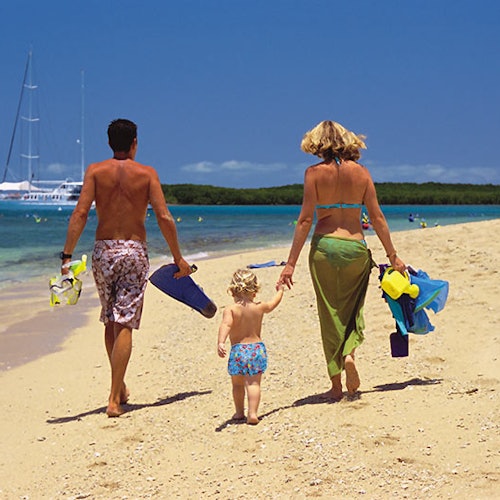 คู่รักและเด็กเดินตามชายหาดทรายไปยังน้ำทะเล โดยถืออุปกรณ์ดำน้ำแบบสน็อกเกิ้ล ในพื้นหลังสามารถมองเห็นเรือใบได้